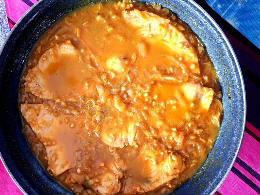 Postas de espadarte a cozinhar num molho espesso de cor laranja‑acastanhada, com pedaços de cebola e alho visíveis.