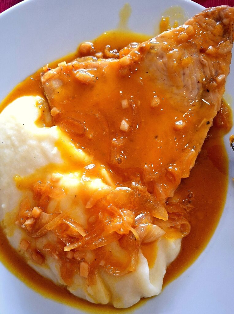 Close-up de posta de espadarte servido sobre puré de batata, coberto com molho espesso e pedaços de cebola.
