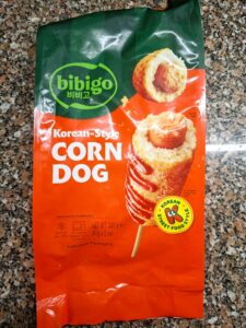Pacote de corn dogs coreanos Bibigo sobre bancada de cozinha, com imagem do produto e cores verde e laranja.