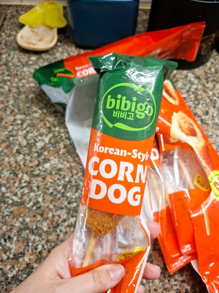 Mão a segurar um corn dog Bibigo ainda dentro da embalagem individual transparente.