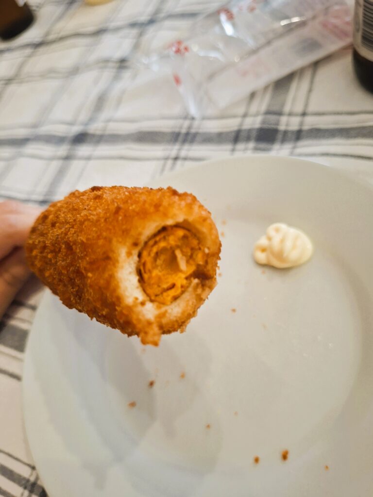 Corn dog with a bite taken out, showing the sausage inside, held above a plate with mayonnaise.