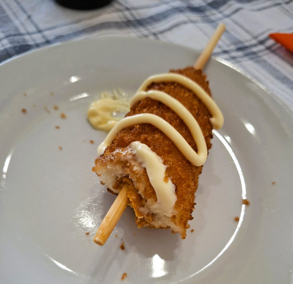 Corn dog com cobertura de maionese, pousado num prato branco.