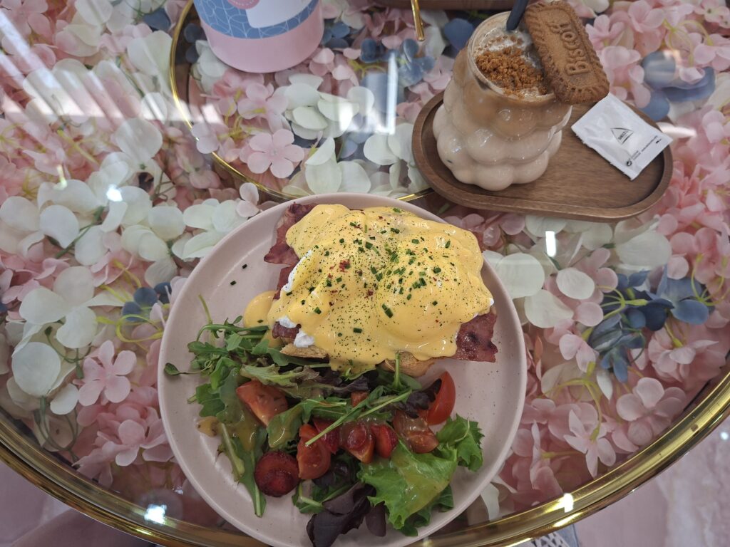 Prato de Brioche Bacon Benedict com salada e latte caramelo servido sobre mesa floral.