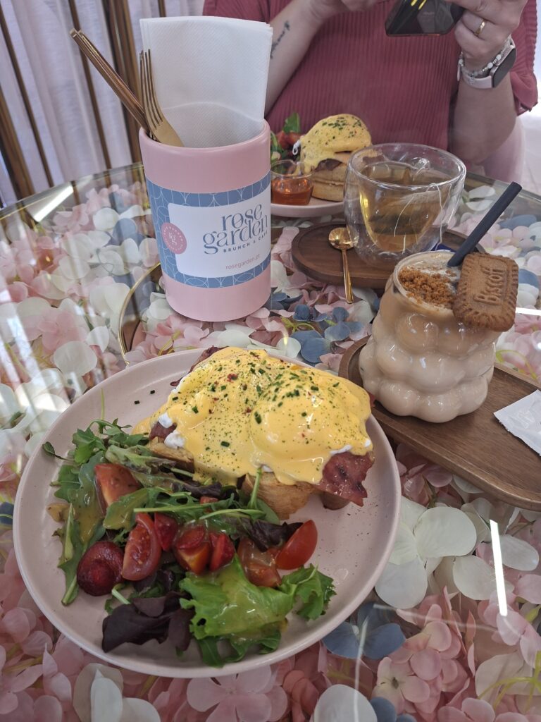 Eggs Benedict, salada, chá e latte com Biscoff sobre mesa decorada com pétalas.