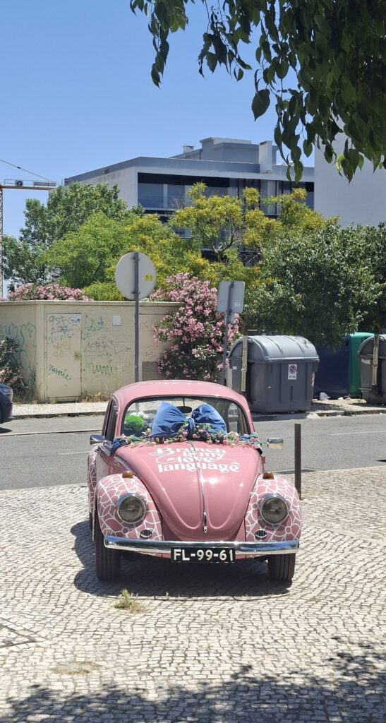 Carro rosa estilo vintage estacionado numa rua, decorado com padrões brancos e laços azuis.