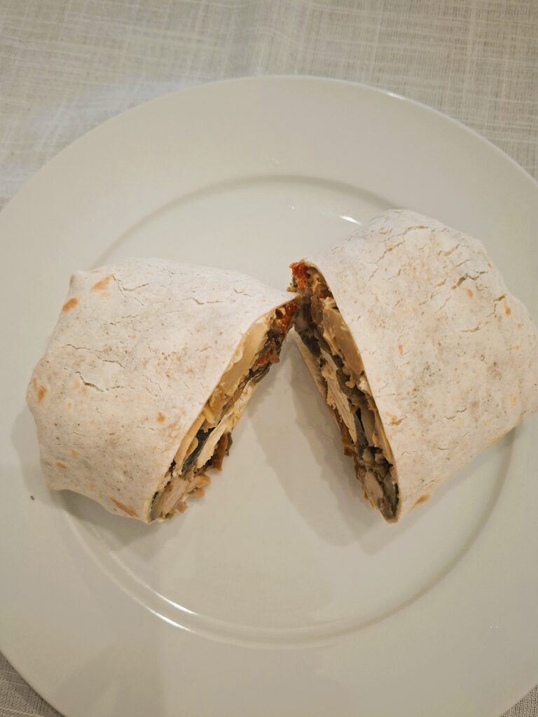 Homemade wrap cut in half, served on a white plate over a light cloth, filled with shredded chicken and roasted vegetables.