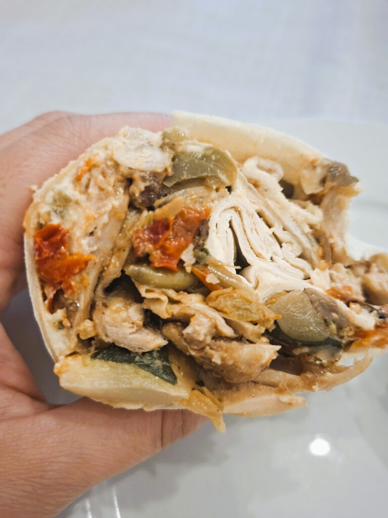 Hand holding half a wrap filled with chicken, roasted vegetables, courgette, mushrooms and sun-dried tomatoes.
