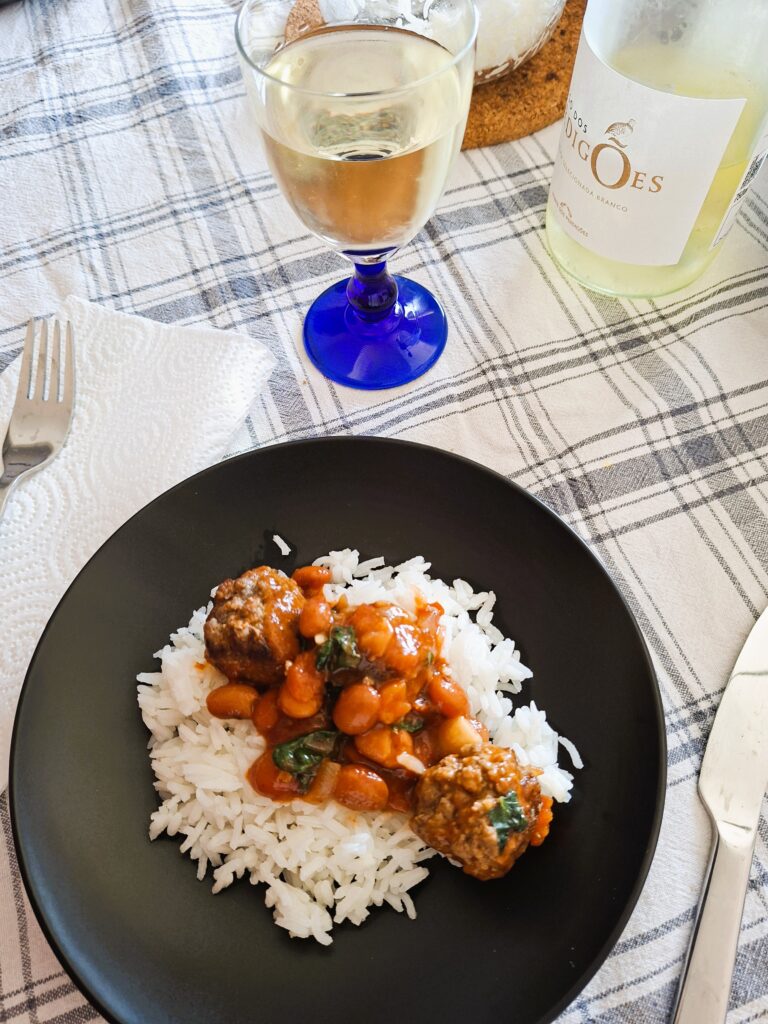 Prato de arroz com almôndegas em molho de tomate com feijão e kale, acompanhado por um copo de vinho branco.