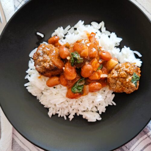 Plate of rice topped with meatballs in tomato sauce with beans and herbs.