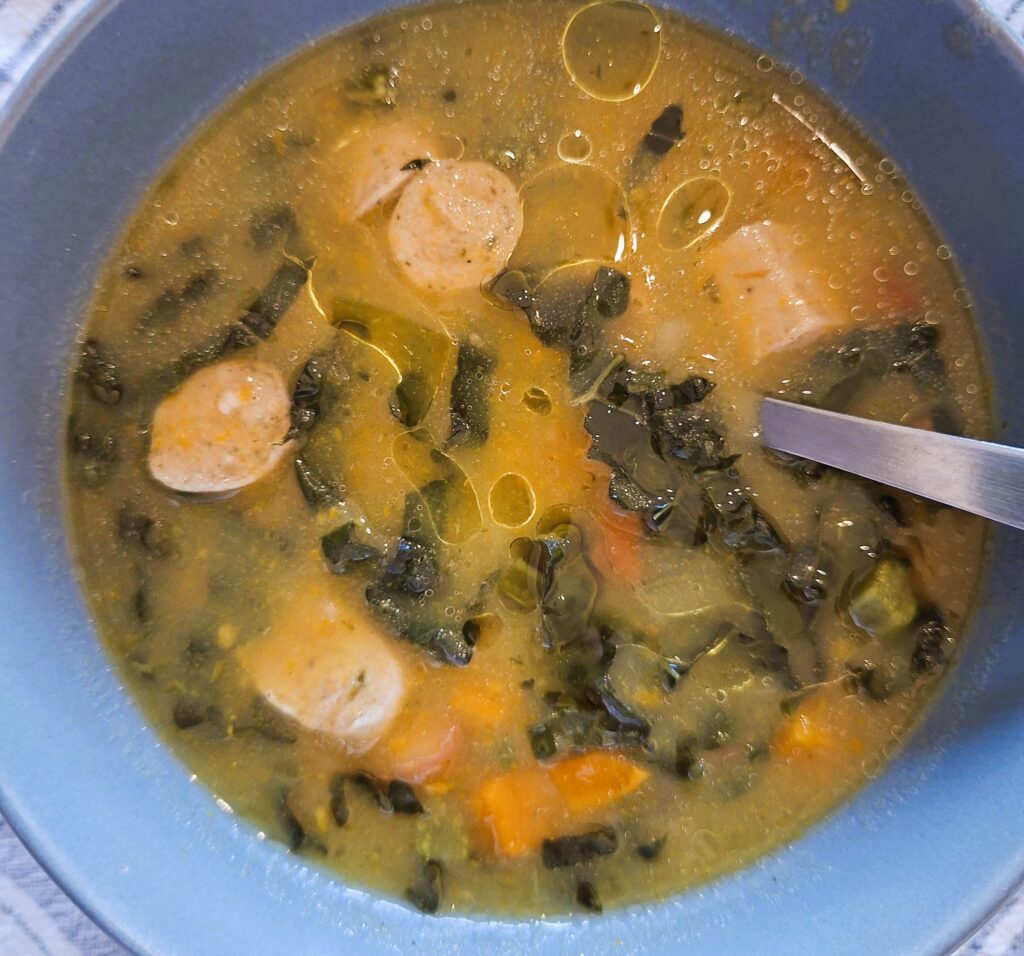 Bowl of homemade soup with sliced sausage, carrots and Tuscan cabbage in a golden broth, with a spoon partially submerged.