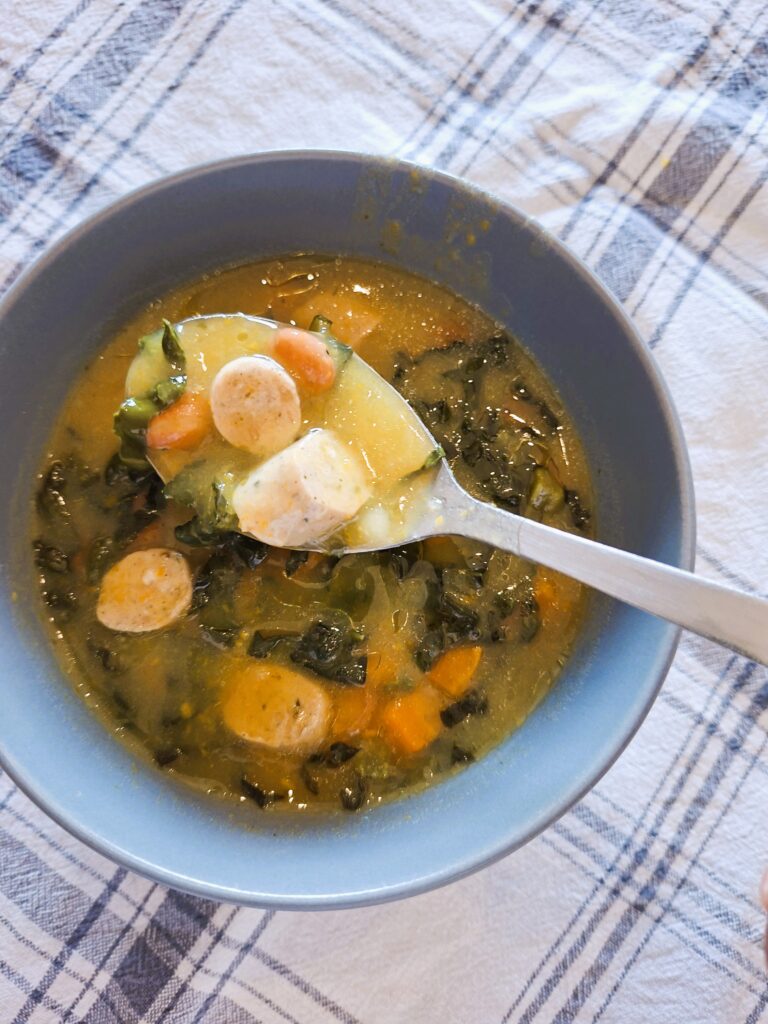 Spoon lifting a portion of creamy soup with sausage, carrots and Tuscan cabbage over a bowl set on a plaid cloth.