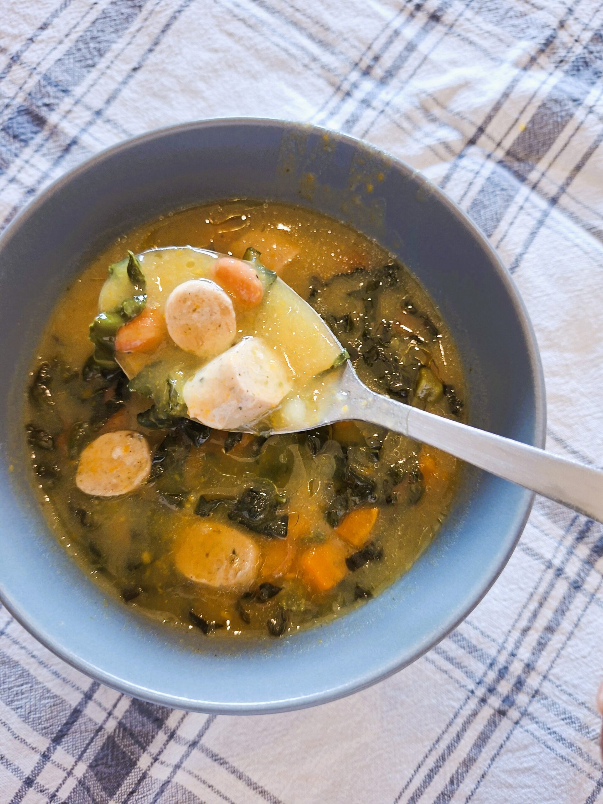 Spoon lifting a portion of creamy soup with sausage, carrots and Tuscan cabbage over a bowl set on a plaid cloth.