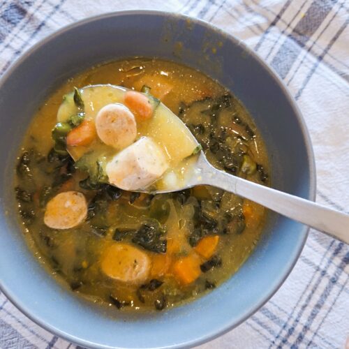 Colher a levantar uma porção de sopa cremosa com salsicha, cenoura e couve toscana sobre uma taça pousada num pano axadrezado.