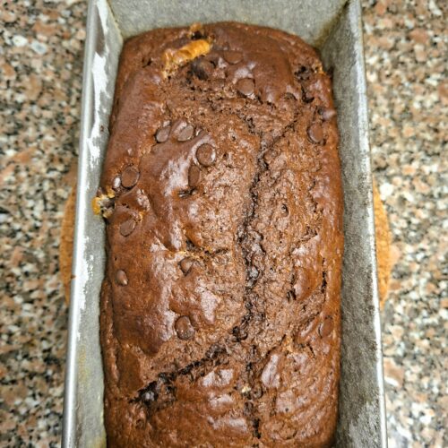 Freshly baked chocolate loaf cake in a metal tin, with a cracked top and melted chocolate chips.
