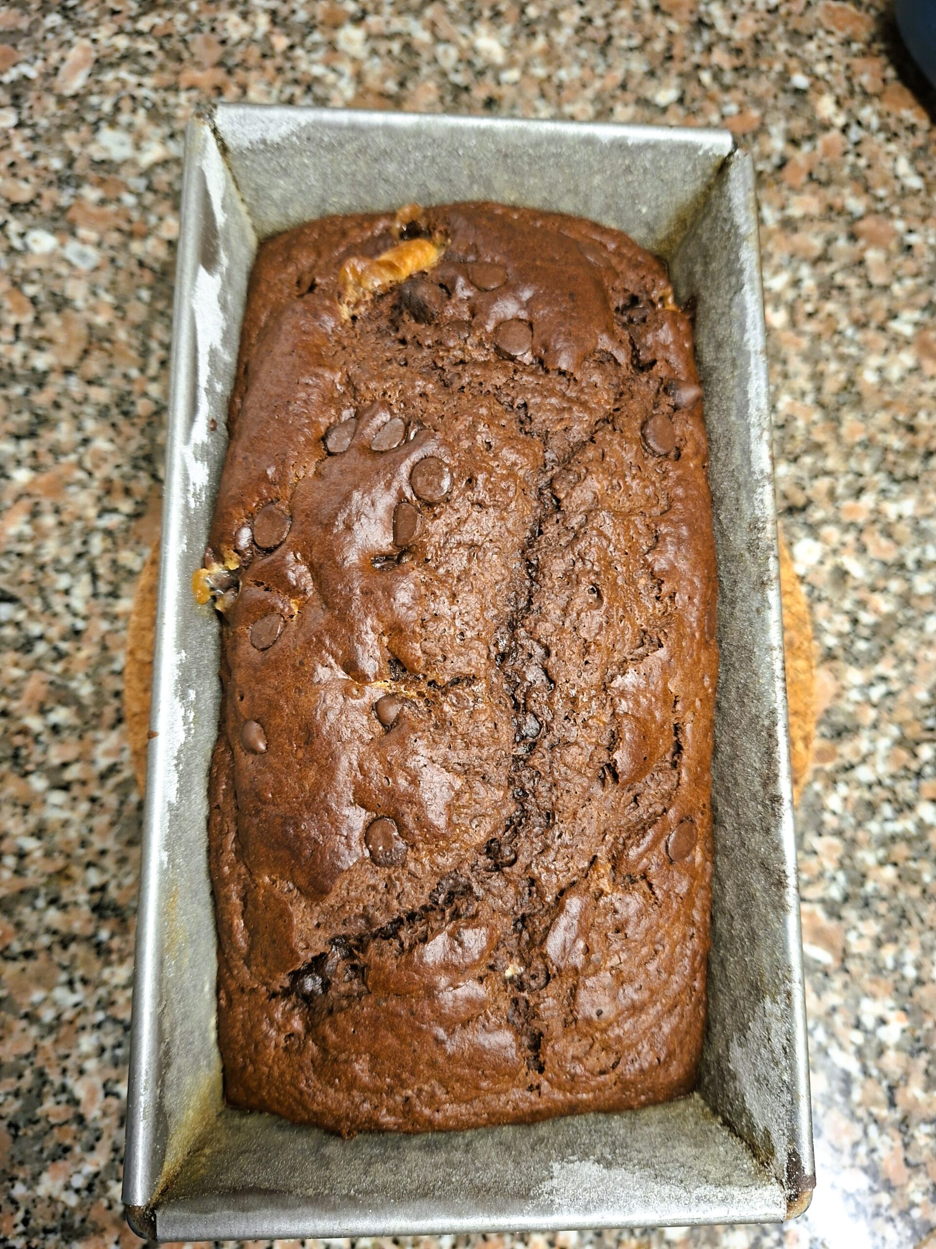 Freshly baked chocolate loaf cake in a metal tin, with a cracked top and melted chocolate chips.