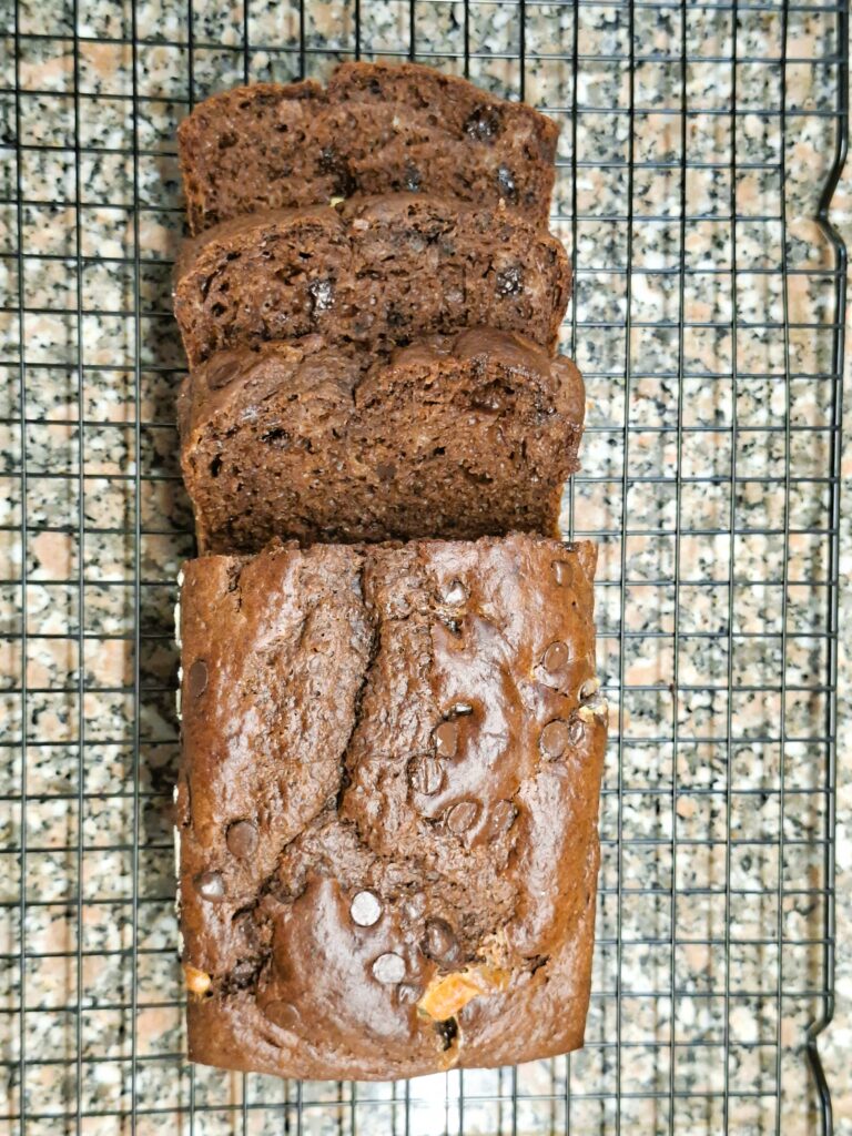 Partially sliced chocolate loaf cooling on a wire rack, with a moist interior and visible chocolate chips.